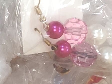 Par de brincos de gancho dourado com uma conta de pérola pink e uma conta facetada rosa, presos numa cartela branca dentro de um saquinho.