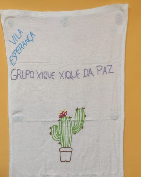 Pano de prato estendido com 'Vila Esperança' e 'Grupo Xique Xique da Paz' bordados em azul e lilás, um cacto florido num vaso e a barra de bolinhas.